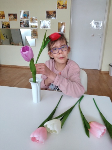 Uczennica siedzi przy stoliku na którym stoi wazon i leżą kwiaty. Dziewczynka ćwiczy wkładanie kwiatów do wazonu.